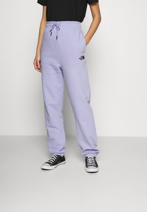 Personne portant un pantalon de survêtement The North Face violet clair avec des poignets élastiques et des poches, associé à des baskets noires et blanches.