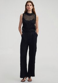 Top nero senza maniche con un motivo a rete e orlo a coste, abbinato a pantaloni neri larghi con texture e décolleté neri con punta aperta.