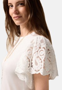 Witte t-shirt met fijne kanten mouwen en een bloemmotief. De stof heeft een zachte textuur, aangevuld met een eenvoudige gouden ketting.