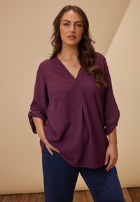 Lila Bluse mit V-Ausschnitt und langen Ärmeln, zwei Brusttaschen und aufgerollten Manschetten; der strukturierte Stoff fällt locker; kombiniert mit blauen Jeans.