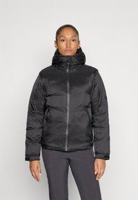 Femme portant une veste rembourrée noire avec un col montant et une capuche, se tenant devant un fond clair et uni.