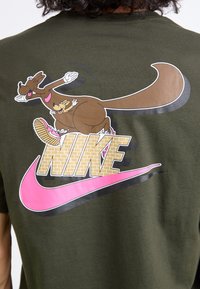Oliven grøn t-shirt med et cartoon-kangaroo grafik, lyserødt Nike-logo og murstensmønster i bogstaverne. Korte ærmer, lavet af bomuld.