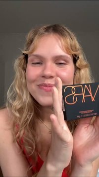 Étui de maquillage compact noir avec des lettres orange, affichant "WAS." Fond flou, mise au point sur le produit tenu par une personne.