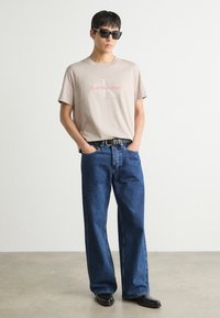 T-shirt en coton beige avec un logo rouge "Calvin Klein Jeans", associé à un jean en denim bleu ample et des chaussures en cuir noir. Modèle portant des lunettes de soleil.