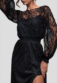 Kvinde iført en sort blonderkjole med gennemsigtige lange ærmer og et højt slid, der holder kjolen ved sliden med den ene hånd.