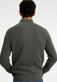 Ribbade, mörkgröna tröja med ståkrage och långa ärmar, som har en avslappnad passform och strukturerad tyg. Bakifrån syns inga tryck.