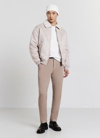 Bomber jacke beige chiaro con colletto in pelliccia, maglietta bianca a girocollo, pantaloni beige e stivali neri alla caviglia. Il modello si trova su uno sfondo neutro.