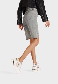 Bermuda shorts di denim blu chiaro con orli sfrangiati, abbinati a sneakers bianche e dorate. Il modello indossa una giacca di pelle nera.