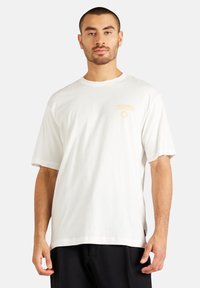 Hvid bomulds T-shirt med afslappet pasform, korte ærmer og et gyldent grafisk design på venstre bryst. Blød tekstur og enkelt design.