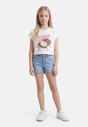 Jeune fille aux longs cheveux blonds porte un t-shirt court blanc avec un motif noix de coco, un short en jean bleu et des baskets blanches, debout droite.