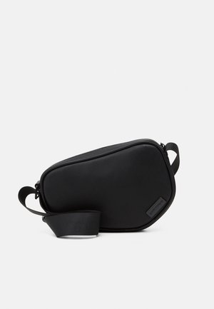 Bolso cruzado negro con cremallera, elaborado en material suave, con una forma plana, una sola correa y un pequeño acento de logo. Diseño minimalista.