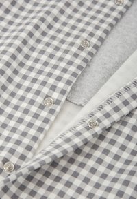 Tela de cuadros en gris y blanco con cierres a presión de metal, superpuesta sobre una base de suave forro polar gris y algodón blanco. Costuras detalladas visibles.