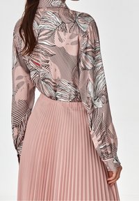 Blusa rosa a maniche lunghe con motivi floreali neri e righe, abbinata a una gonna rosa plissettata, che mette in mostra texture morbide e linee pulite.