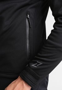 Chaqueta de forro polar negra con un bolsillo con cremallera, puños con orificio para el pulgar y un logo en el antebrazo, hecha de un material suave y texturizado.