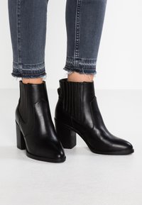 Bottes courtes en cuir noir avec un talon épais, dotées de panneaux latéraux élastiques et de détails côtelés pour une finition texturée.