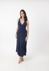 Mørkeblå ærmeløs kjole med V-udskæring, tætsiddende overdel og fløjtende nederdel. Modellen bærer lyse beige sandaler med stropper. Glat stofstruktur.