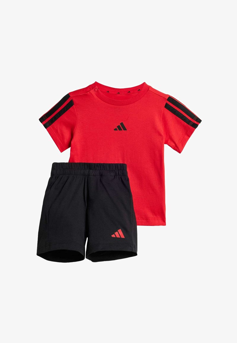 Crvena pamučna majica s crnim prugama na ramenima i logotipom, uparena s crnim kratkim hlačama koje imaju mali crveni logotip. Elastični pojas na kratkim hlačama.