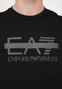 Black cotton t-shirt featuring a gray logo design with "EA" and "Emporio Armani" text, highlighting a smooth texture and crew neck style.