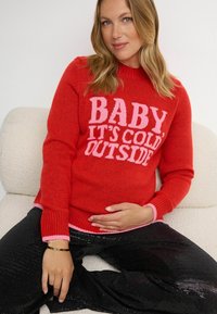Czerwony sweter z dzianiny z różowym napisem "KOSTKA, JEST ZIMNO NA ZEWNĄTRZ", w połączeniu z czarnymi spodniami z cekinami, z kontrastującymi różowymi mankietami.