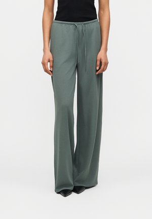 Femme portant un pantalon vert taille haute à jambes larges avec un cordon de serrage, un haut noir et des chaussures pointues noires, debout devant un fond uni.