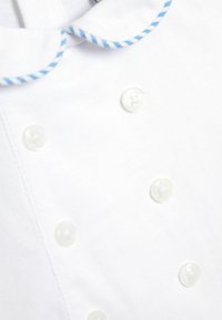 Vêtement en tissu blanc avec un col rond, des bords rayés bleus et plusieurs boutons blancs présentant une surface claire et texturée.