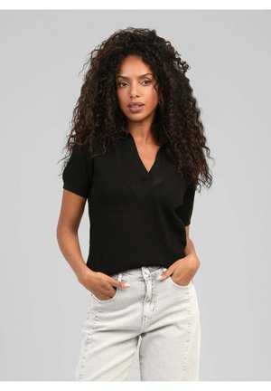 Femme aux cheveux bouclés portant un haut en maille noir à manches courtes et un jean clair taille haute, mains dans les poches, sur un fond gris uni.