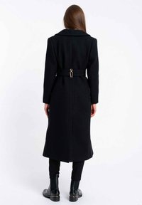 Długi czarny płaszcz z kołnierzem, przewiązany w talii paskiem z metalicznym akcentem, o prostym kroju. Noszony na czarnych spodniach i botkach za kostkę.