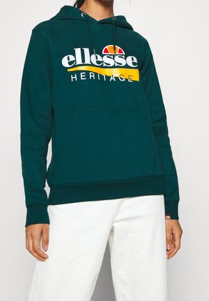 Ciemnozielona bluza z kapturem ellesse Heritage z logo, noszona przez osobę w białych spodniach, widoczna górna część ciała i ręce, bez pokazania głowy.