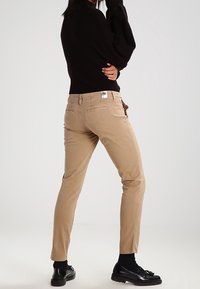 Pantalons beige slim fit avec poches arrière et patch logo. Portés avec un pull noir et des chaussures, présentant une texture en coton lisse.