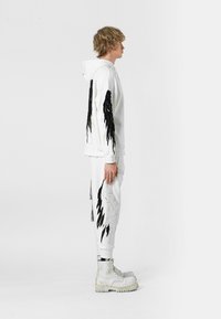 Sudadera blanca y pantalones con motivos de alas negras. Botas de goma. Detalles texturizados en las alas, creando un contraste dinámico contra el blanco.