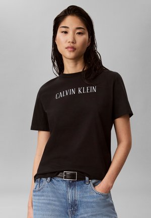 Ung kvinde med vådt mørkt hår bærer sort Calvin Klein t-shirt og blå jeans med hænderne i lommerne mod en ensartet grå baggrund.