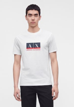 Junger Mann trägt ein weißes Armani Exchange T-Shirt mit schwarzem und rotem Logo, steht vor einem einfarbig hellen Hintergrund.