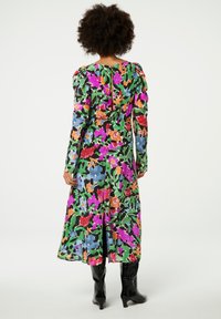 Bloemige jurk met lange mouwen, voorzien van een levendig, veelkleurig patroon op een groene achtergrond, een getailleerde taille en een rits aan de achterkant. Gestyled met zwarte laarzen.