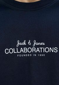 Laadukkaasta puuvillasta valmistettu laivastonsininen t-paita, jossa on pyöreä kaula-aukko. Paidassa on valkoisella painettuna teksti: "Jack & Jones", "KOLLABORATIOT" ja "PERUSTETTU 1990".