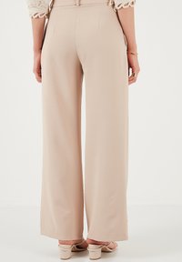 Beige wide-leg trousers with a smooth texture, featuring a high waist and back pockets. Styled with pale block-heeled sandals.