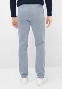 Man die lichtblauwe broek met achterzakken en witte sneakers draagt, staand tegen een effen witte achtergrond.