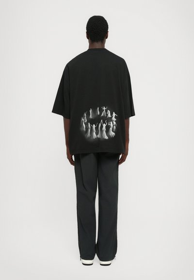 Person facing away wearing black oversized t-shirt with a white circular ghostly figure design on the back, paired with black pants and white shoes.