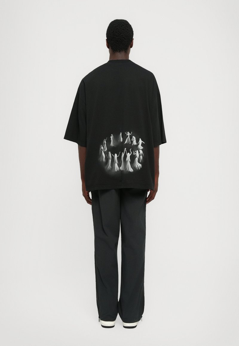 Persoon met de rug naar de camera, gekleed in een zwart oversized t-shirt met een witte cirkelvormige spookachtige figuur op de rug, gecombineerd met zwarte broek en witte schoenen.
