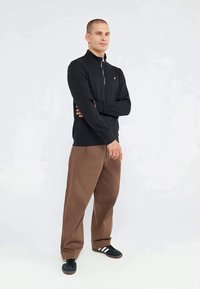 Man standing with arms crossed wearing black zip-up sweater, brown loose pants, and black sneakers with white stripes on white background.