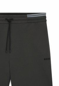 I pantaloni della tuta grigio scuro presentano un cinturino con coulisse, due strisce nere nella parte superiore e un logo nero vicino all'orlo, realizzati in tessuto morbido.