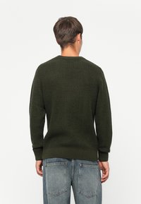 Un maglione verde scuro con un design a maglia texturizzata, scollatura rotonda e polsini a costine, abbinato a jeans blu sbiaditi.