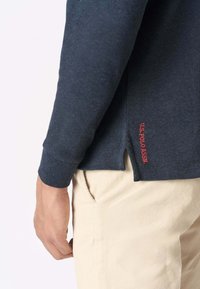 Camicia a maniche lunghe blu navy con spacco laterale, texture morbida e logo rosso "U.S. Polo Assn." all'orlo, abbinata a pantaloni color crema.