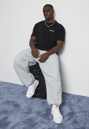 Black short-sleeve t-shirt with white logo, gray baggy pants, and white sneakers. Smooth fabric, simple design, seated on a black drawer unit.