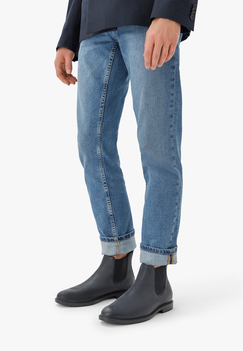 Melni ādas čelija zābaki ar elastīgām sānu paneļiem, apvienoti ar gaiši zilu džinsu biksēm ar sarullētām piedurknēm un vieglu izbalējumu.