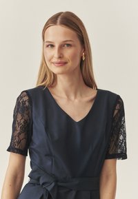 Tumesinine kleit, millel on V-kaelus, lühikesed pitsist varrukad ja kangast vöö. Silmapaistev tekstuur ja voolava disainiga elegantne ning keha järgnev siluett.