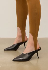 Black leather mule heels with pointed toes and slim stiletto heels, paired with brown fitted legging featuring cut-out details.