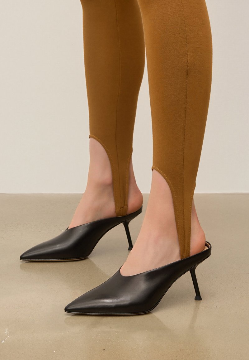 Black leather mule heels with pointed toes and slim stiletto heels, paired with brown fitted legging featuring cut-out details.