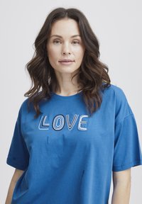 Camiseta de algodón azul con un gran texto bordado "LOVE" en múltiples colores. Mangas cortas y corte relajado, con ligeras manchas de agua visibles.