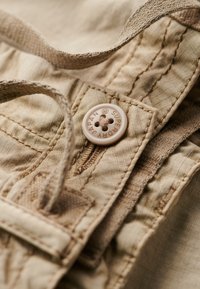 Beige stof met een getextureerde afwerking en stiksel details. Een ronde, lichtbruine knoop met de woorden "Super Japan" en een bloemmotief.