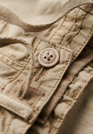 Gros plan sur un tissu beige présentant une surface texturée, un bouton rond portant l'inscription "SUPER JAPAN" et des détails brodés le long des bords.